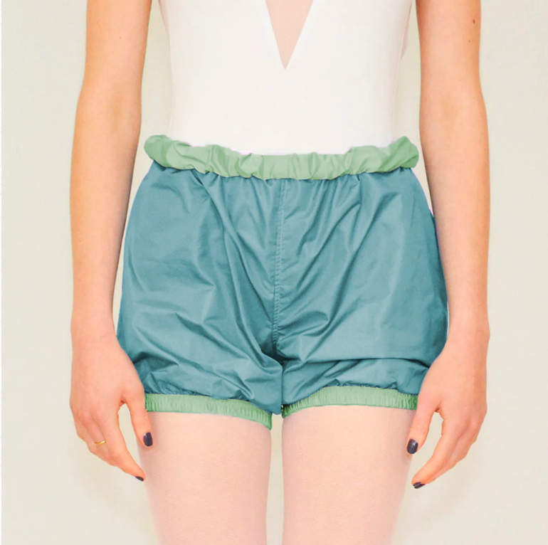 A dancer wears teal trash-bag style shorts with a sage green waistband.