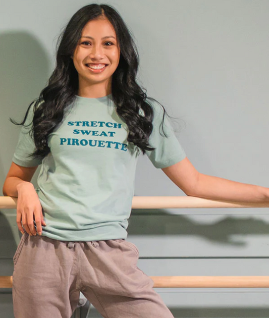 A dancer leans against a ballet barre wearing a sage green t shirt with "STRETCH SWEAT PIROUETTE" written down the center in darker green